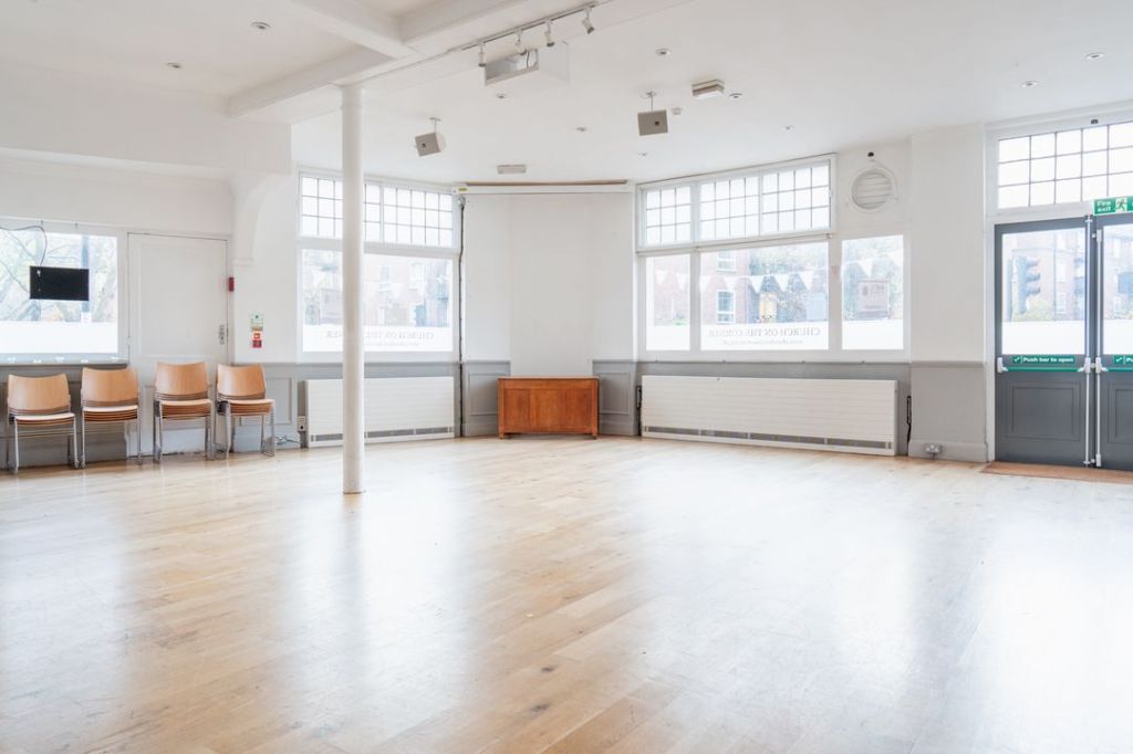 Picture of our main hall, with wooden floors, white walls and large windows, and chairs stacked up underneath a window.